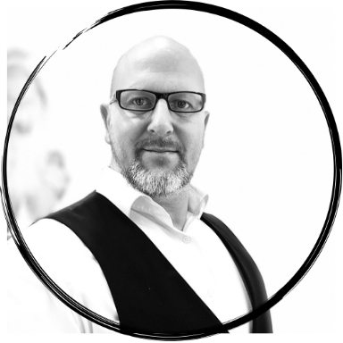 Mann in schwarzem Weste und Brille vor weißem Hintergrund, lächelnd.