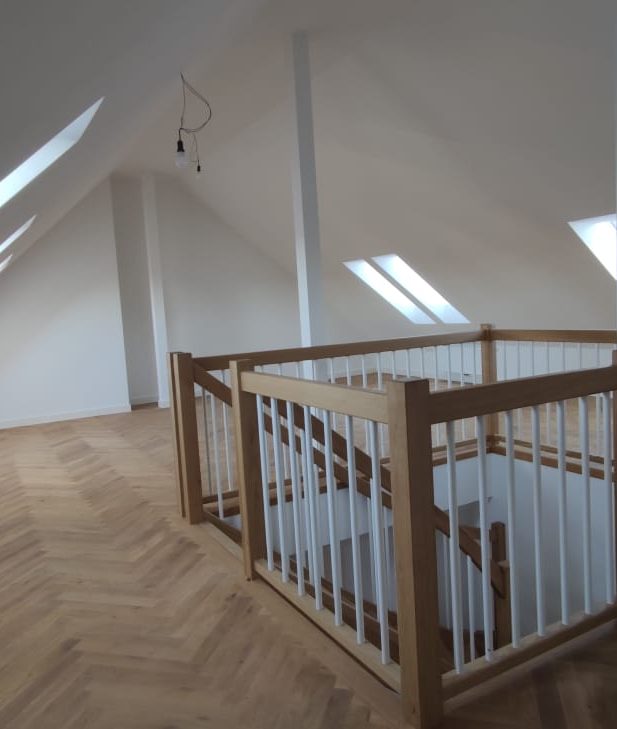 Heller, leerer Raum im Dachgeschoß mit Holztreppe und Geländer aus Holz.